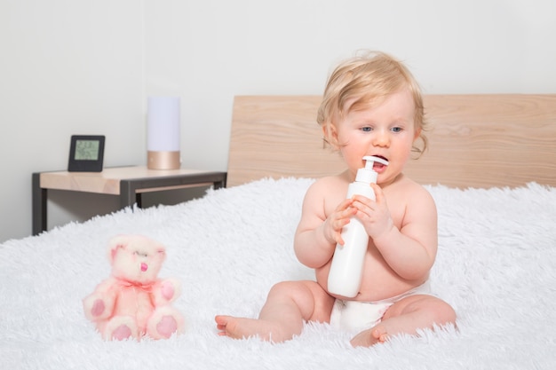 Cute little baby girl with bottle of baby oil in parent bedroom.