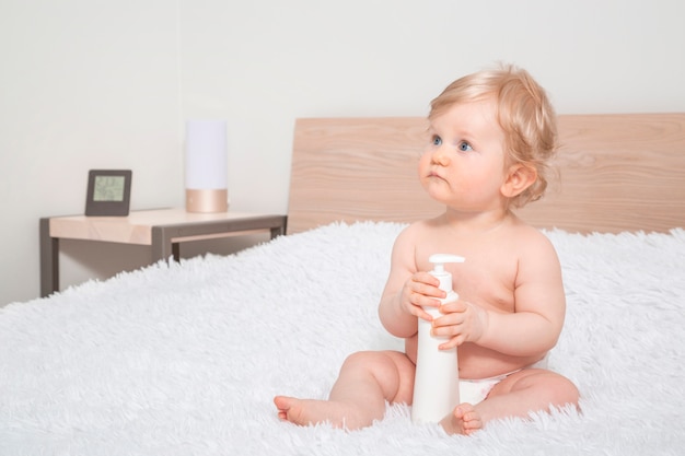 Cute little baby girl with bottle of baby oil in parent bedroom.