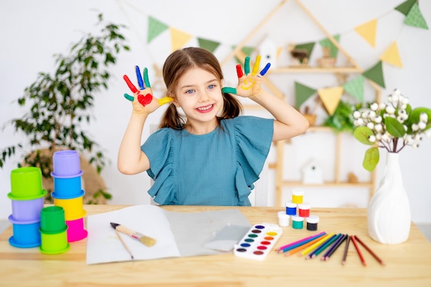 美しい小さな赤ちゃんの女の子がペンとペンで絵を描くことを学ぶ幼稚園の指の塗料で彩られた手で色の多い絵を描いています