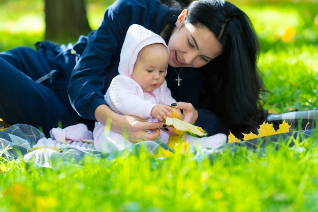 Cute little baby playing with an autumn leaf as her mother cuddles with her on a blanket in the park