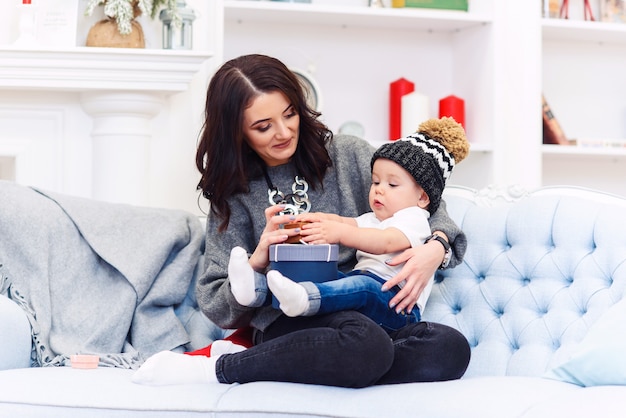 축제 장식 된 크리스마스 룸에서 편안한 블루 소파에 앉아 그녀의 어머니의 무릎에 휴식하는 귀여운 작은 아기.
