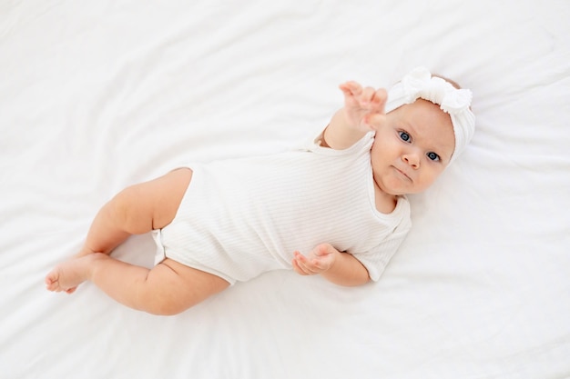 귀여운 작은 아기가 집에 있는 흰색 침대에 바디슈트를 입고 누워서 웃고 있는 갓난 아기의 윗면을 보고 있습니다