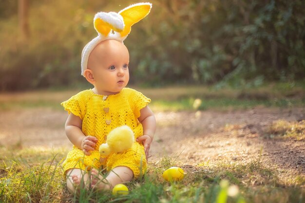 イースターの日にバニーの耳を持つかわいい赤ちゃん 女の子は芝生の上でイースターエッグを探します 夕日の光線の中でイースターエッグとチキンを持つ女の子