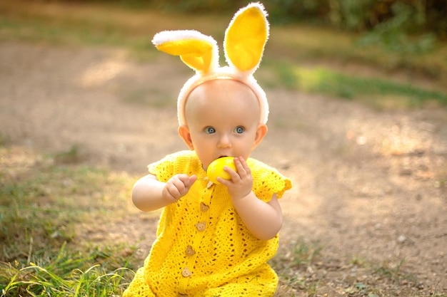 イースターの日にバニーの耳を持つかわいい赤ちゃん 女の子は芝生の上でイースターエッグを探します 夕日の光線の中でイースターエッグを持つ女の子