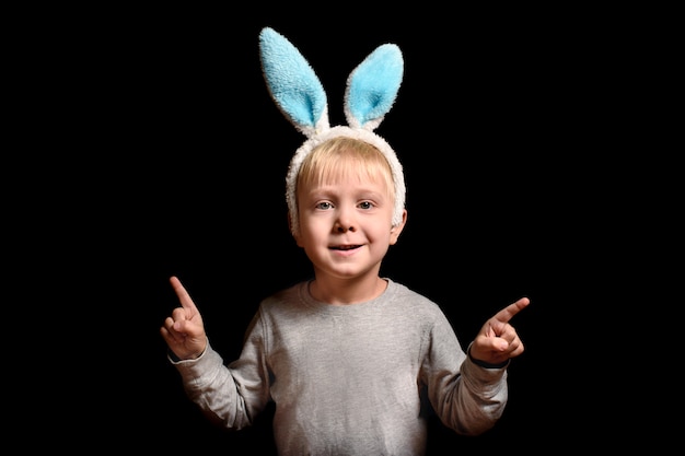 Cute little blond boy in hare ears shows his index fingers upwards stands on black 