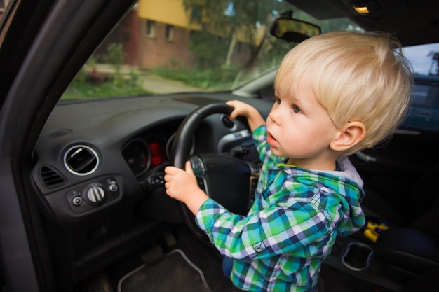 The cute little blond boy is sitting behind the wheel of a modern car and looking away