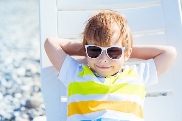 Cute little blond boy in sunglasses sits on a sunbed on the ocean coast