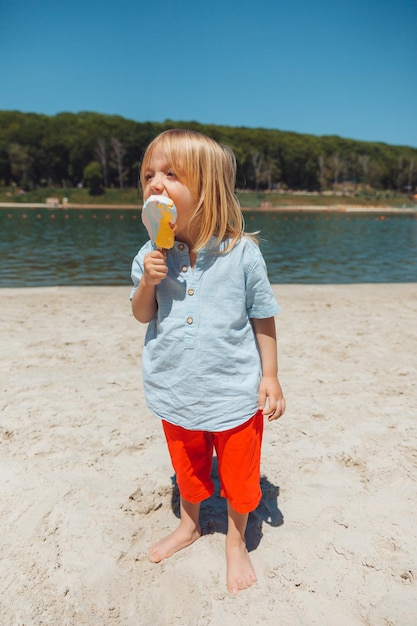 Симпатичный маленький блондин с длинными волосами ест мороженое на пляжном отдыхе