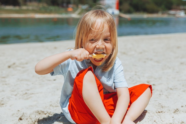 Cute little blond boy with long hair eating ice cream on the beachsummer vacation