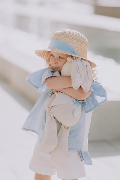 Carina bambina bionda in abito blu e cappello di paglia che gioca vicino alla fontana con coniglietto di peluche, carta