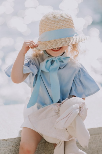 Cute little blond girl in blue dress and straw hat playing near\
fountain with plush bunny, card