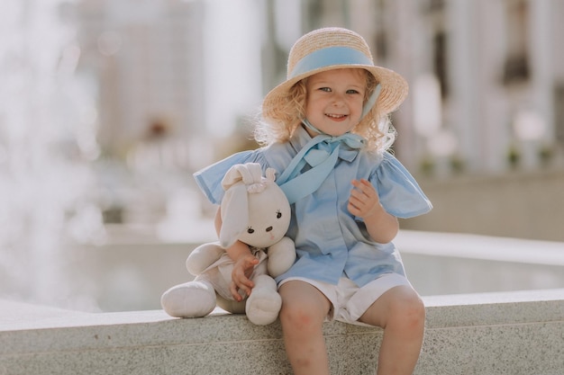 cute little blond girl in a straw hat and a blue dress plays with a stuffed rabbit outdoors