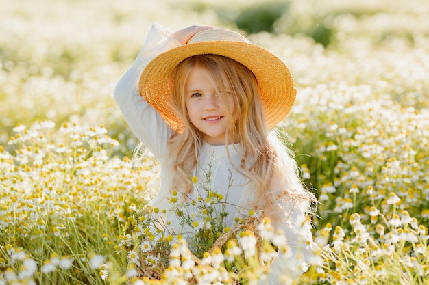 Cute little blonde girl in a cotton dress and straw hat walks in a field of daisies collects them in the basket