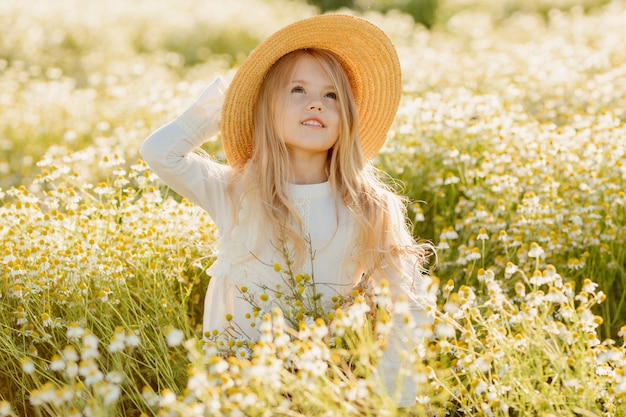 Cute little blonde girl in a cotton dress and straw hat walks in a field of daisies collects them in the basket