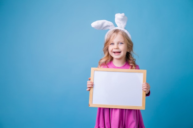 Cute little blonde girl in the ears of an Easter bunny holding a white empty board for announcements.