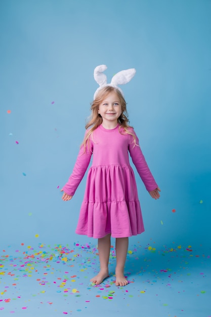 青い背景の上のピンクのドレスのイースターバニーの耳のかわいい小さなブロンドの女の子。