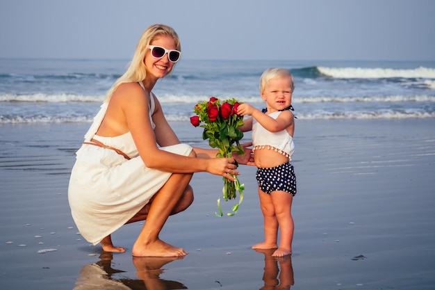 Photo cute little blonde girl giving mom a bouquet of roses morning on the beach by the sea.mother's day and birthday celebration.valentine's day february 14 beautiful woman and her daughter ocean vacation