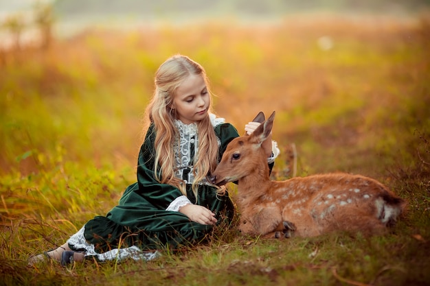 Una ragazza bionda carina in un abito vintage verde si siede accanto a un piccolo cervo sika intorno a loro bellissimi alberi gialli autunnali.