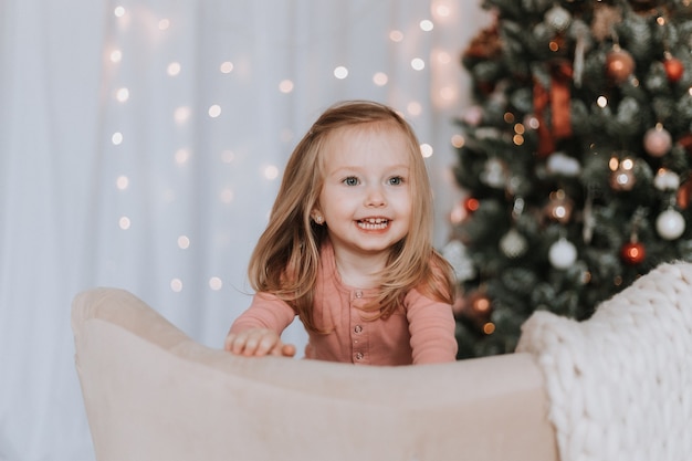 귀여운 금발 소녀가 크리스마스 트리 배경의 의자에 무릎을 꿇고 있다