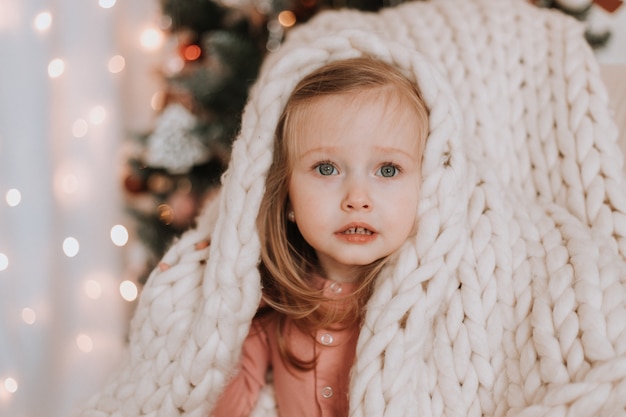 cute little blonde girl is sitting wrapped in light plaid of large knitting near the Christmas tree