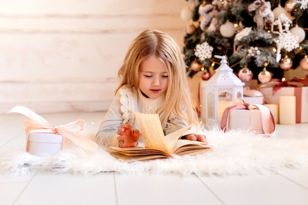 Cute little blonde girl reads a book at home near the Christmas tree