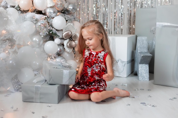 cute little blonde girl in a red dress with gifts near the Christmas tree photo zone