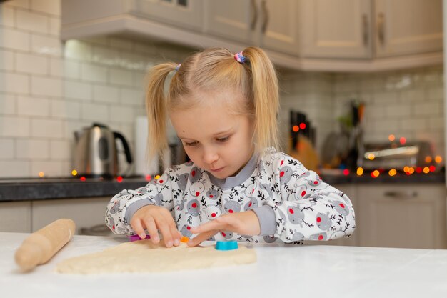 カラフルな形のかわいいブロンドの女の子は、生地からクッキーを切り取ります小さな料理人は準備します