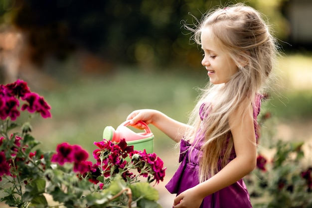 Cute little blonde girl with long hair watering flowers in summer pelargonium geranium child smiling planting flowers in sunny garden Little girl gardener Family with children work in the backyard