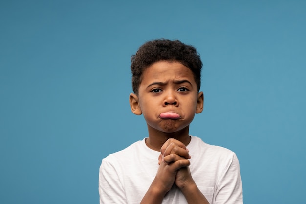 Cute little boy of African ethnicity holding his hands by chin with pleading expression while standing on blue