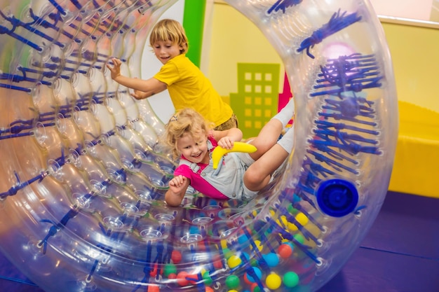 Фото Симпатичный маленький мальчик и девочка, играющие в zorb - катящееся пластиковое цилиндрическое кольцо с отверстием посередине в помещении