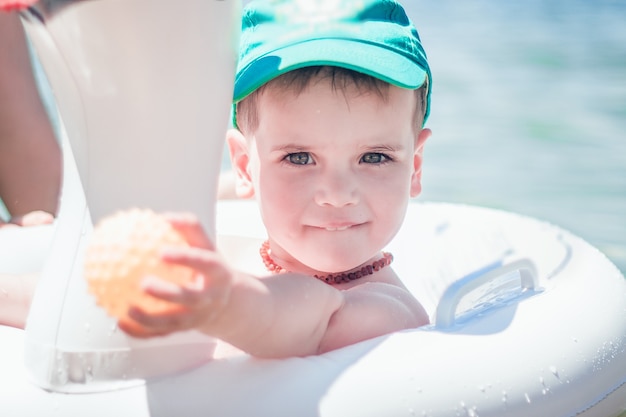 A cute little boy in a blue baseball hat floating in the sea in an inflatable ring.