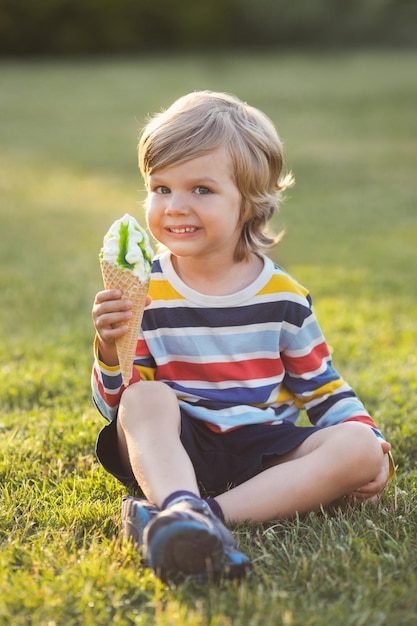 Cute little boy eating ice cream and sitting on green grass in park at summer day