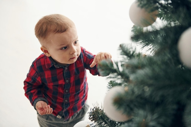 Cute little boy in festive clothes is indoors near christmas tree at New year time