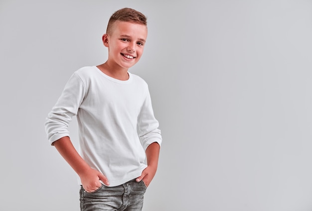 Cute little boy on a gray background holds his hands in his jeans pockets and smiles