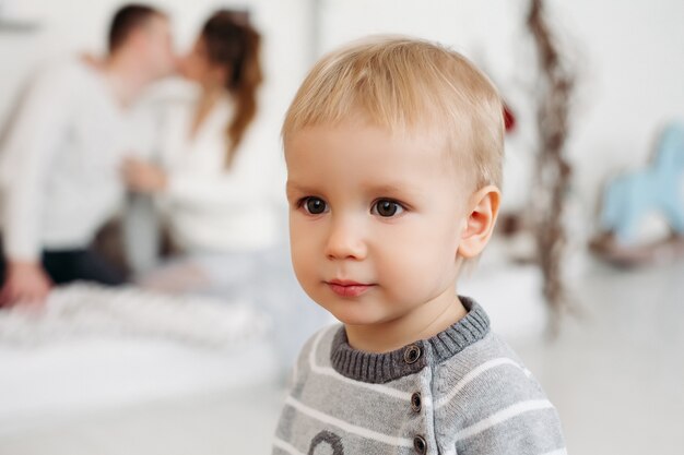 Cute little boy in gray sweater standing in studio and looking away while young parents in love hugging and kissing behind