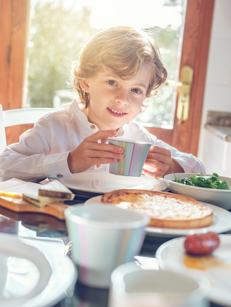 Cute little boy having breakfast and sitting at table while smiling and looking at camera in dining room