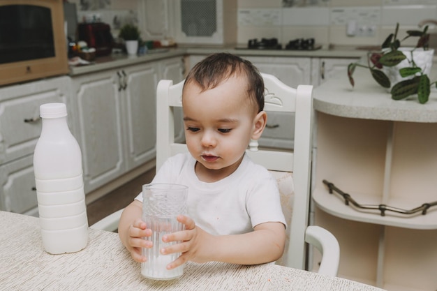 Il ragazzino sveglio sta bevendo il latte al tavolo nel mocap della bottiglia di latte della cucina