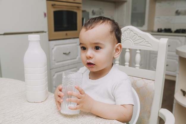 Il ragazzino sveglio sta bevendo il latte al tavolo nel mocap della bottiglia di latte della cucina