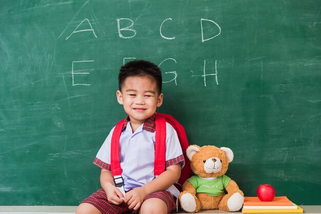 Cute little boy kindergarten preschool in student uniform with school bag sit with teddy bear on green school blackboard