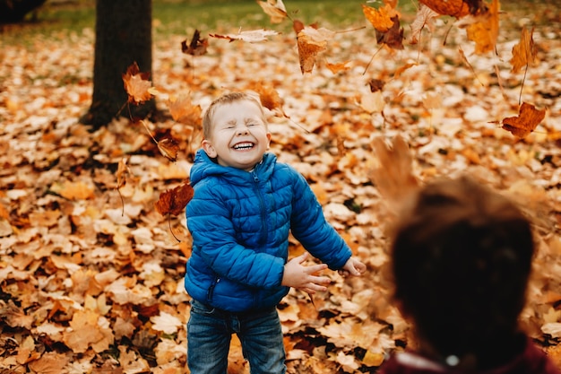 Cute little boy laughing and having fun while playing with the leaves in the park with her mother.