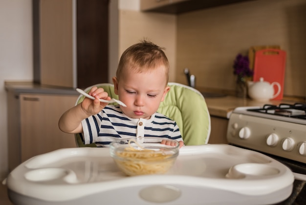 Фото Милый маленький мальчик учится есть сам с вилкой. ребенок ест спагетти, сидя на высоком стуле на кухне.