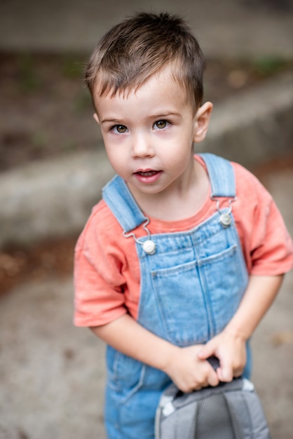 a cute little boy looks at the camera holds a satchel in his hands dressed in denim overalls