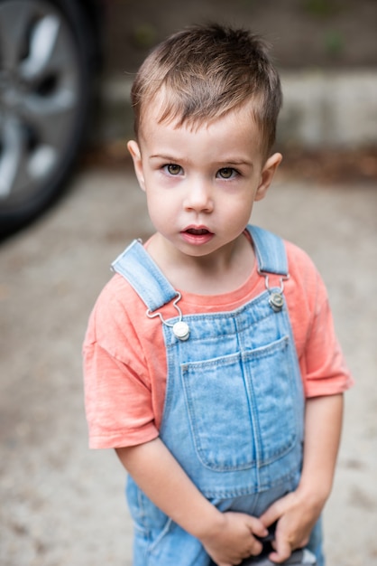 A cute little boy looks at the camera holds a satchel in his hands dressed in denim overalls