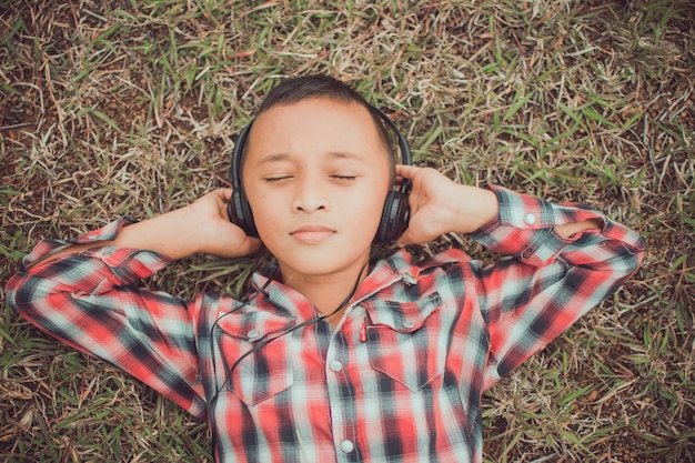 Cute little boy lying on grass with headphone for listening in the park, her face feel like happy with sunshine. subject is blurry.