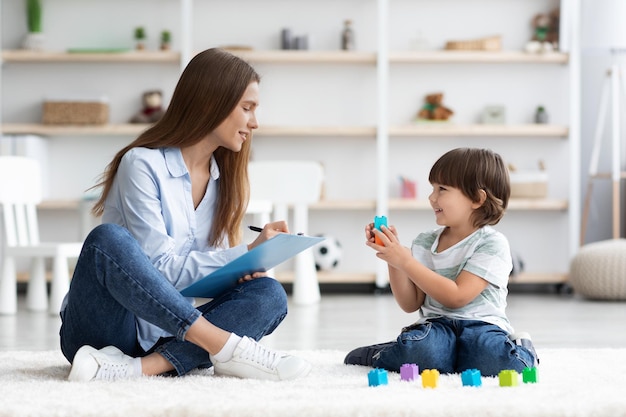 Cute little boy playing with colorful blocks young professional woman kids psychologist writing