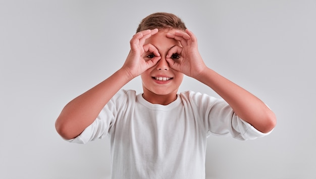 Foto ritratto di un ragazzino carino su sfondo grigio, con le dita incrociate come occhiali sugli occhi e sorridente.