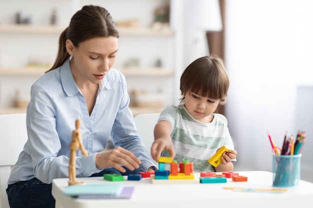 Cute little boy preschooler playing developing wooden games with professional woman psychologist at