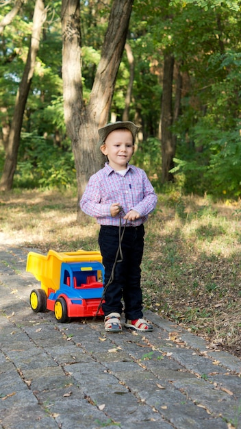 森の舗装された車線で彼の後ろにカラフルなおもちゃのトラックを引っ張るかわいい男の子