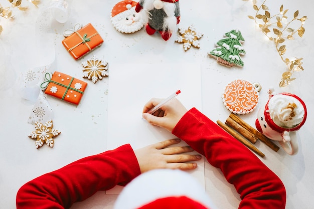 Cute little boy in red sweater and Santa hat writes letter to Santa Claus asking to bring presents top view New Year and Christmas concept DIY child kid believes in miracles