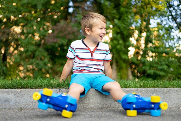 A cute little boy in roller skates is sitting on the grass
smiling looking at the camera the concept of sports outdoor games
active games